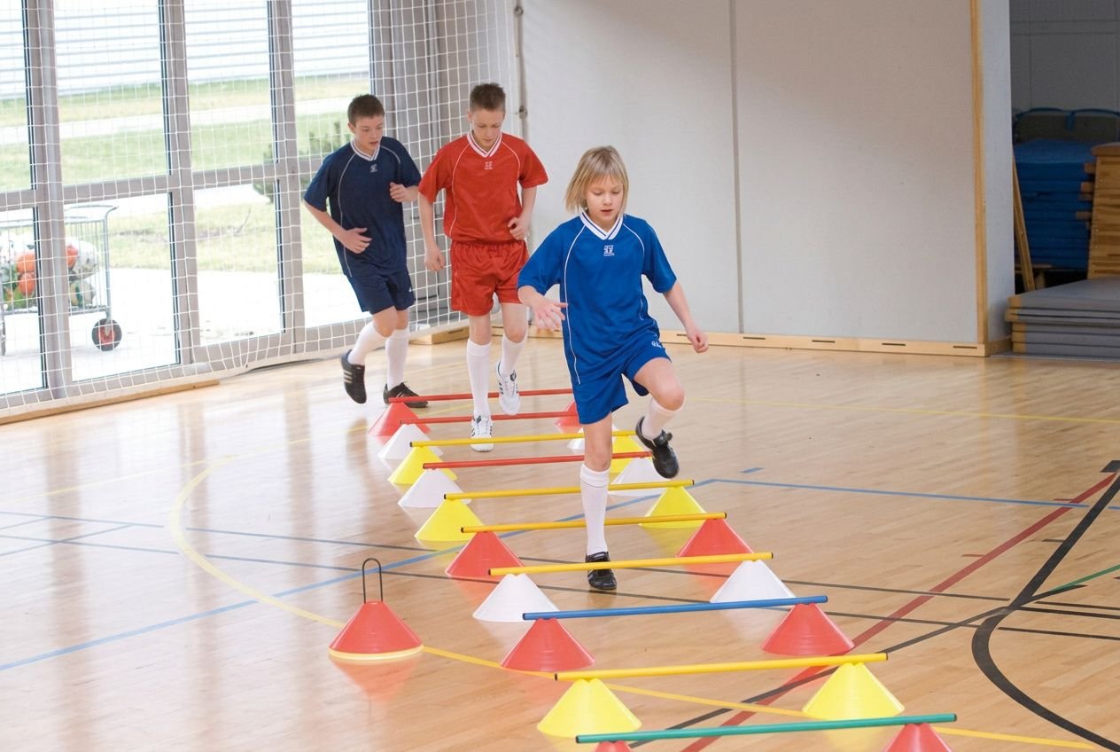Kinderen in een gymzaal oefenen met hindernissen: springen over pylonen en balken, met kleurrijke ringen en kegels op de vloer. Gymmateriaal basisschool in gebruik.